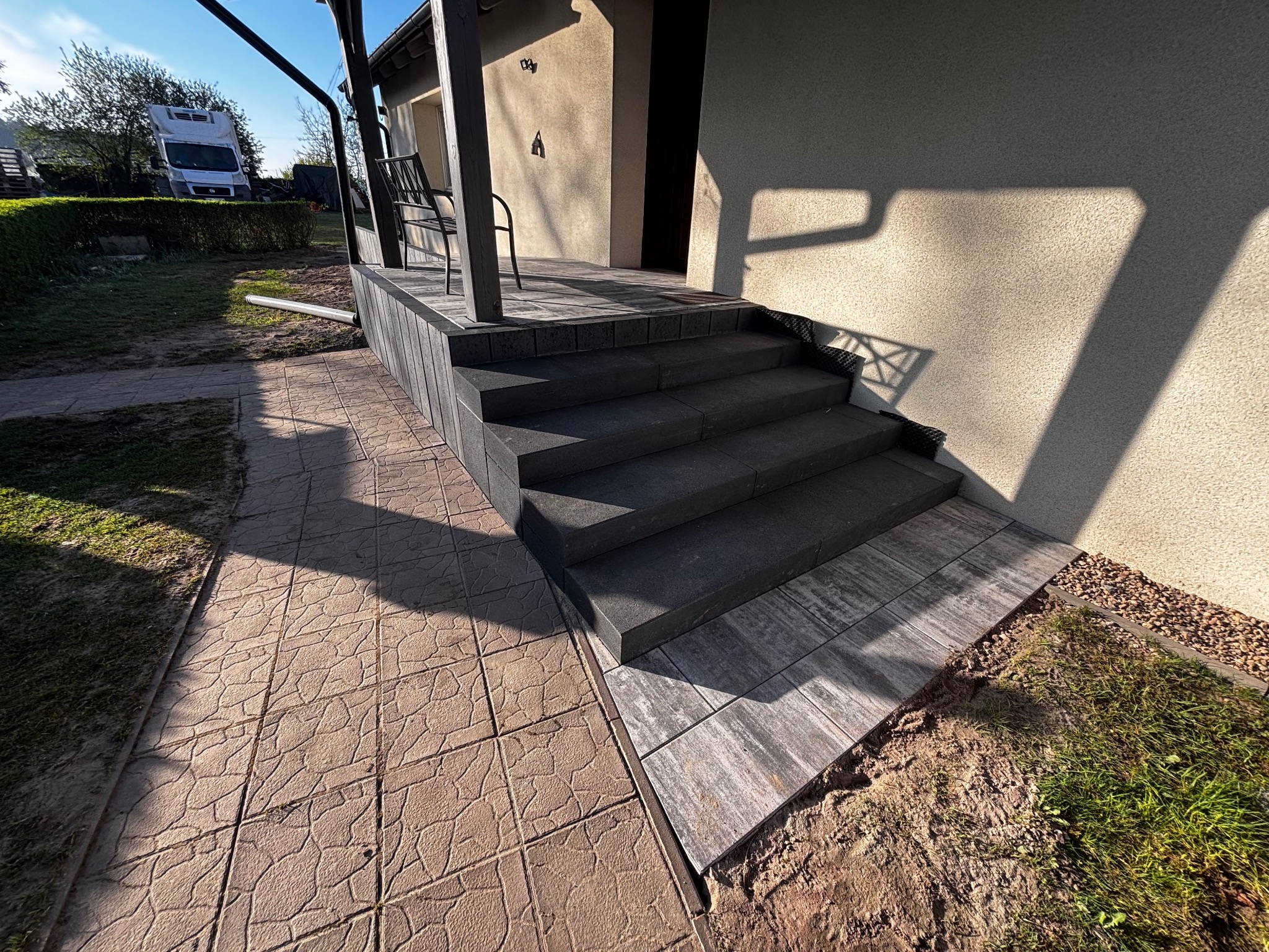 Schody z kostki brukowej prowadzące do domu, z widocznym fragmentem chodnika i trawnika. Detale wykończenia, oświetlenie słoneczne.