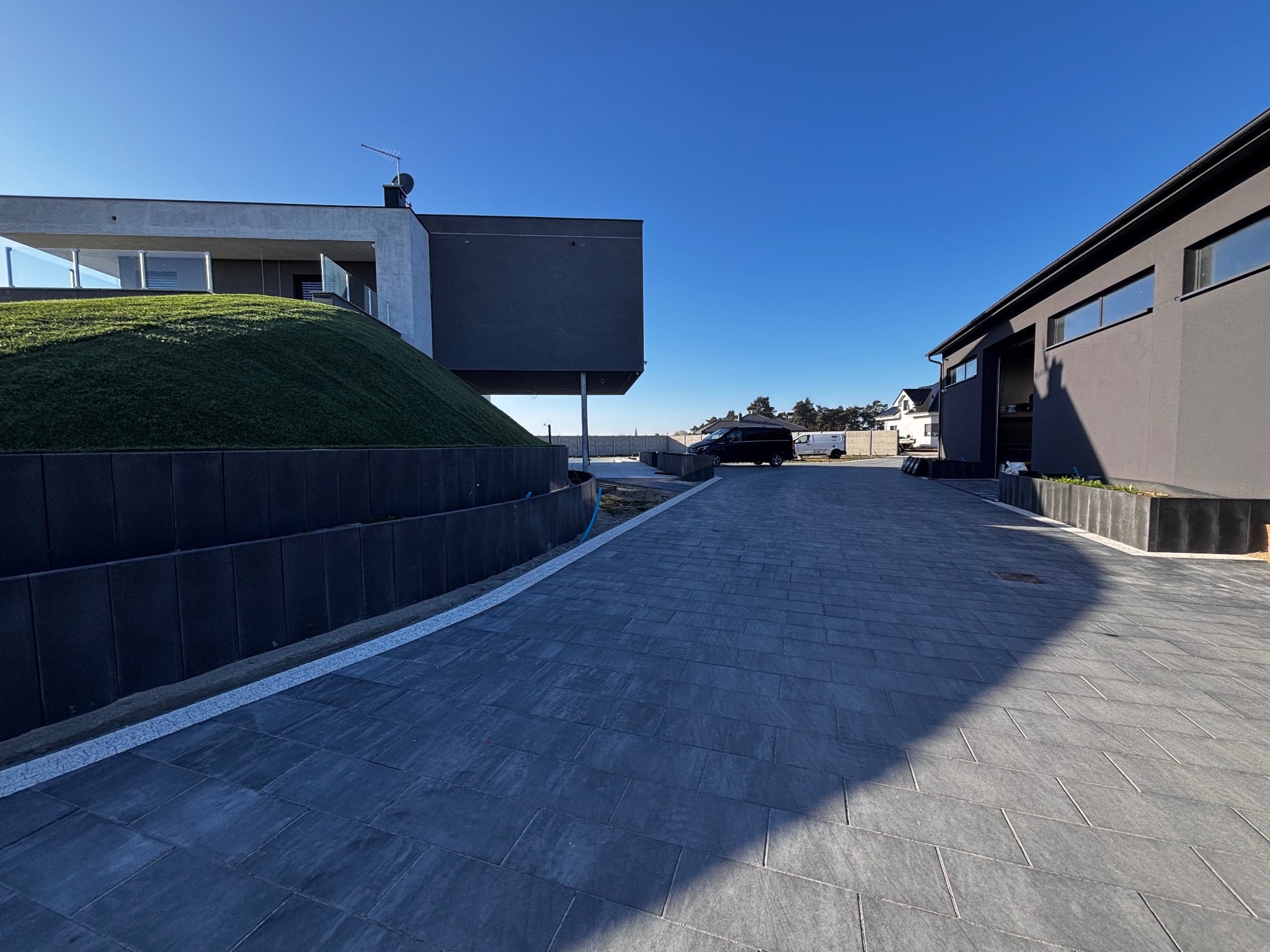 Nowoczesna posesja z ciemnoszarą kostką brukową, elegancko wkomponowaną w architekturę budynków. Jasne fugi podkreślają kształt kostki. Błękitne niebo w tle.