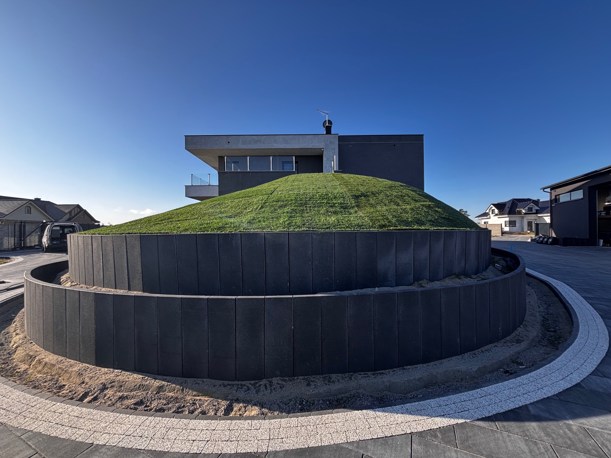 Nowoczesna posesja z okrągłym podwyższeniem z kostki i trawy, otoczona brukowanym chodnikiem. Minimalistyczny dom w tle pod błękitnym niebem.