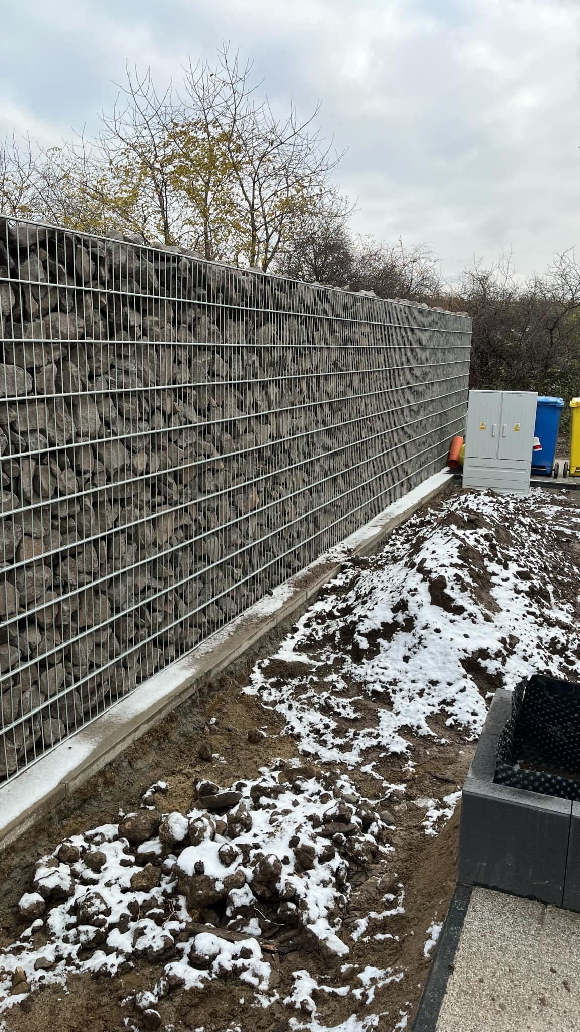 Gabionowa ściana z kamienia w metalowej siatce, częściowo pokryta śniegiem. W tle widoczne drzewa i szafka elektryczna. Elementy małej architektury.