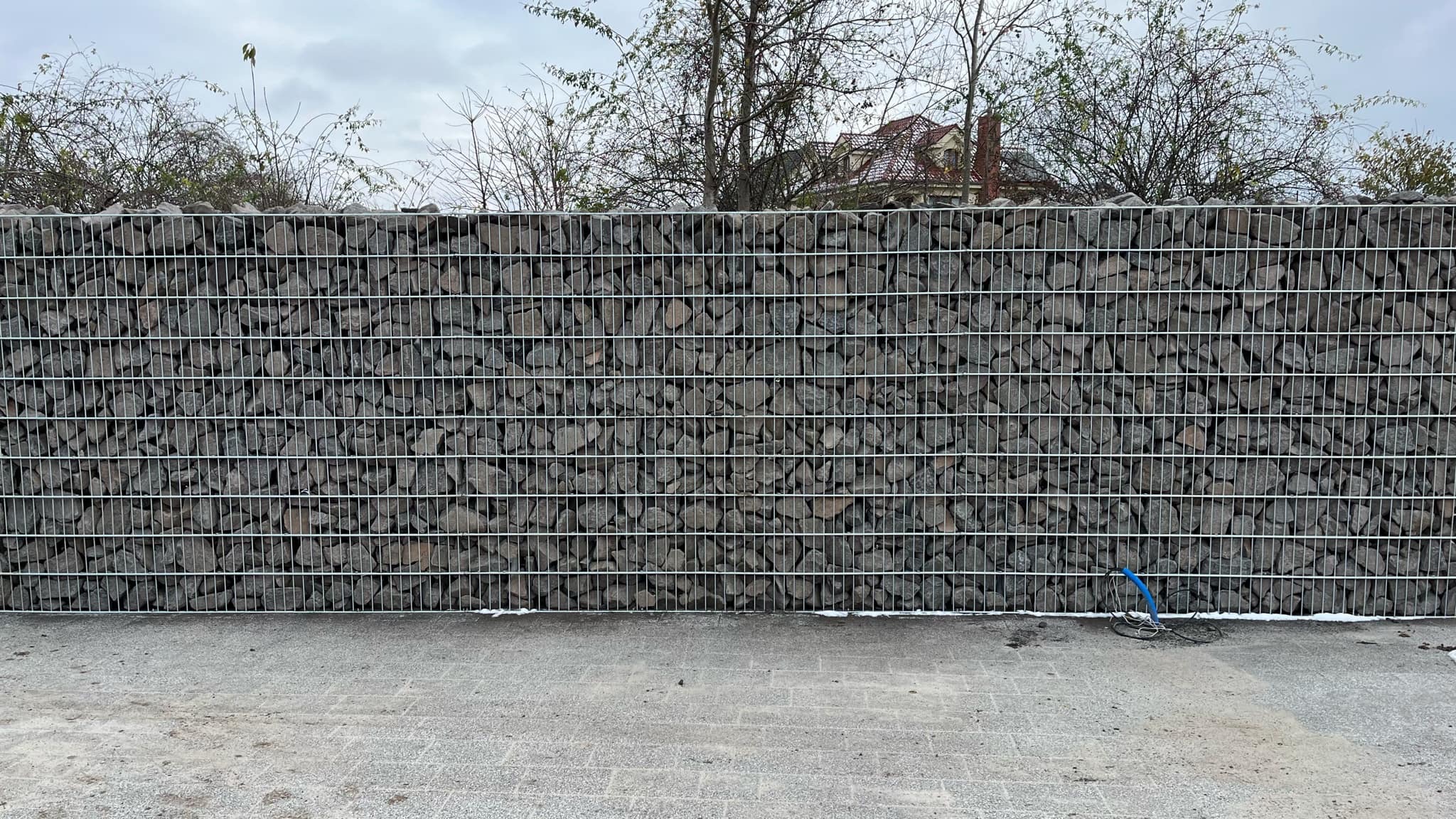 Gabion z szarego kamienia w metalowej siatce tworzy ogrodzenie. Widoczne drzewa i dach domu w tle. Na dole kostka brukowa i zwój niebieskiego kabla.
