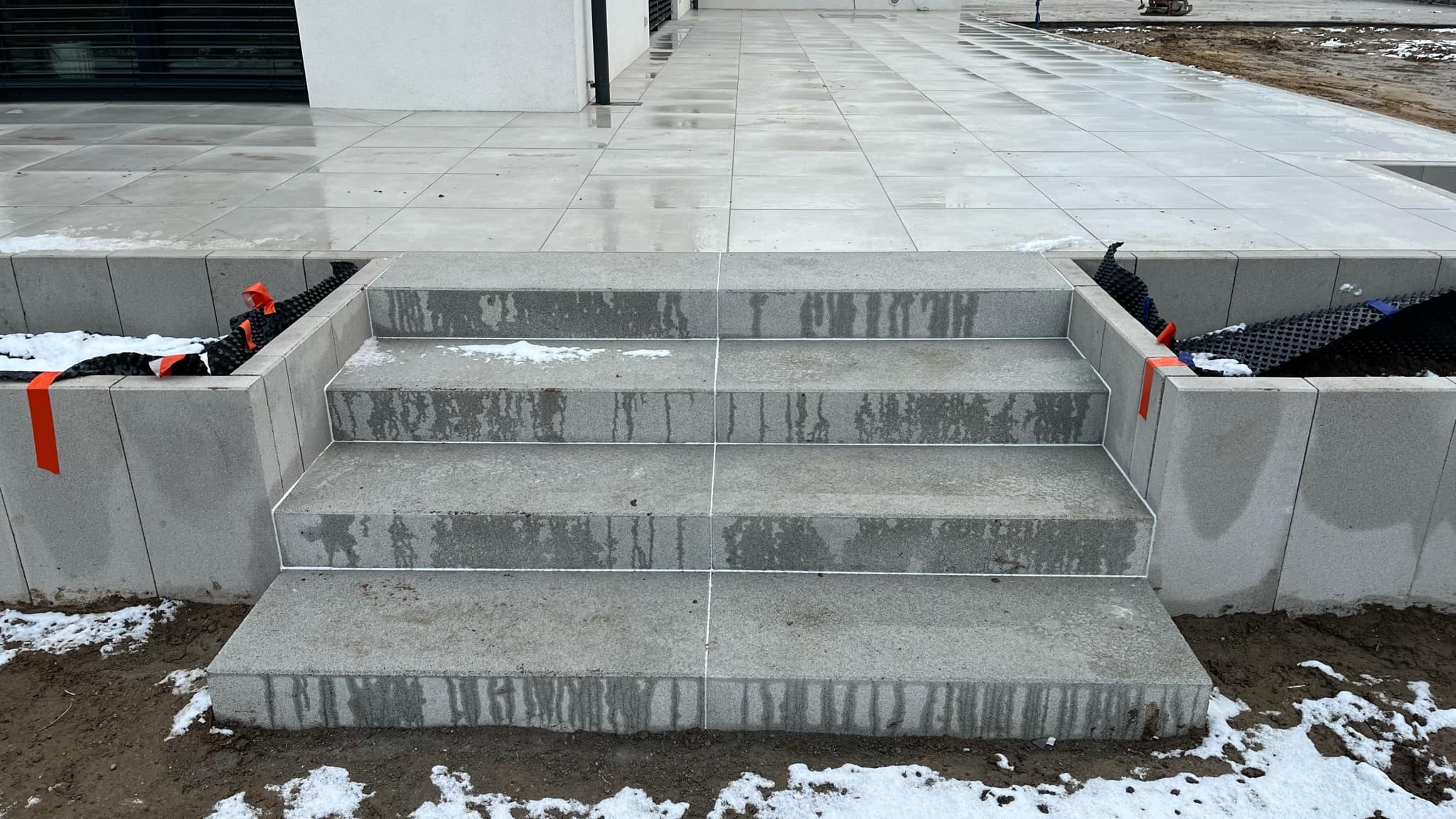 Schody z szarych płyt betonowych z widocznymi fugami, obok wyłożony taras z jasnych płyt, na ziemi resztki śniegu. W tle biały budynek.