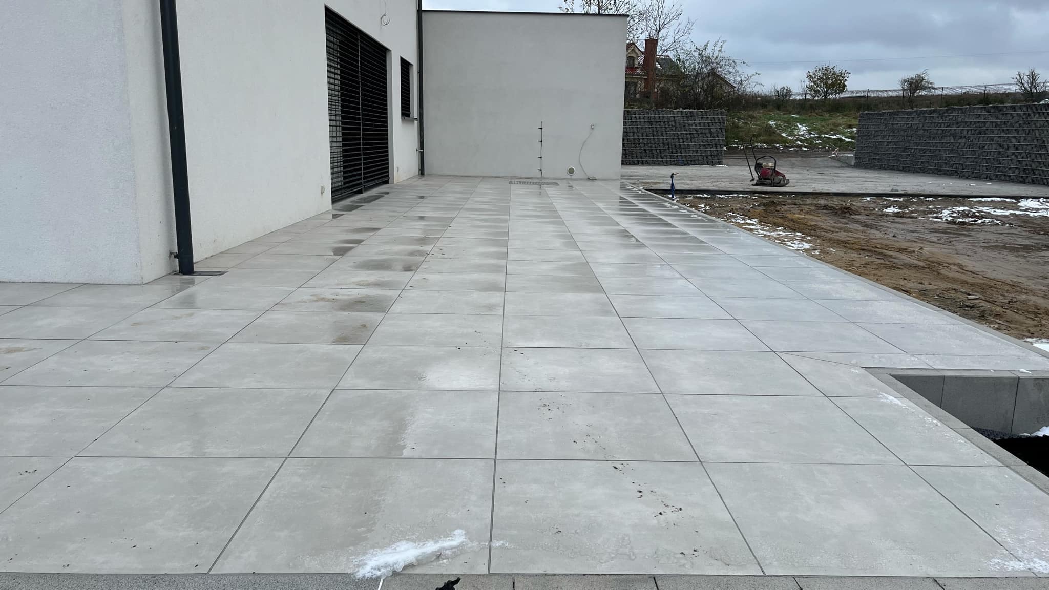 Nowoczesny taras wyłożony jasnoszarą, kwadratową kostką. Widoczne świeże ślady po deszczu, dom z białymi ścianami i fragment ogrodu w tle.