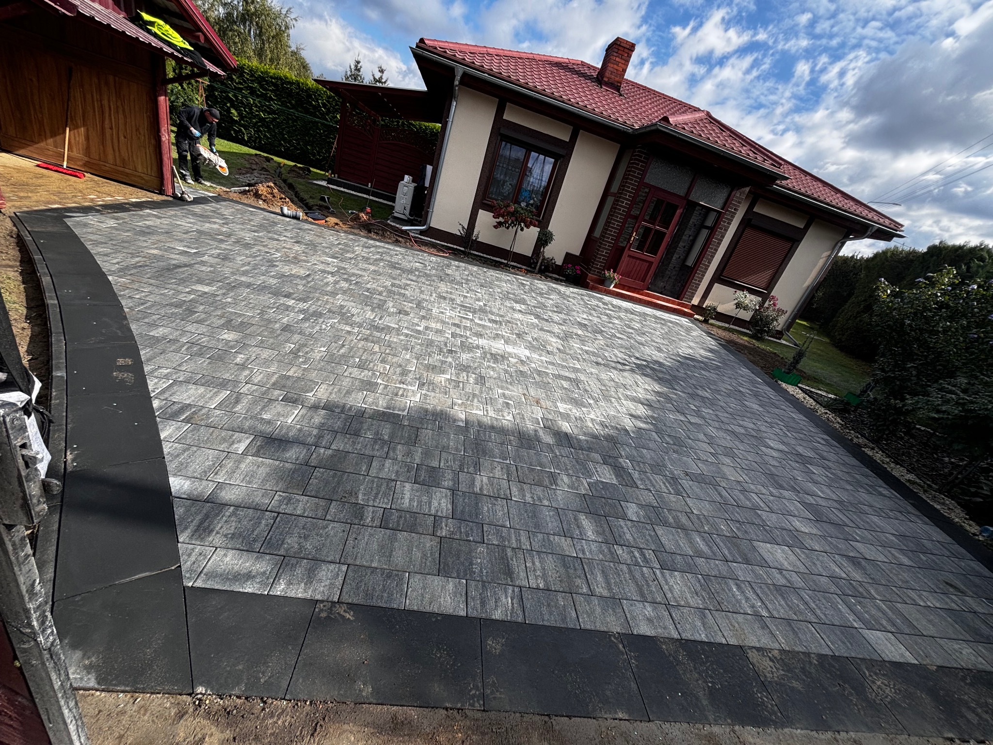 Ułożona szara kostka brukowa na podjeździe przed domem z czerwoną dachówką i pracownikiem w tle, widok z perspektywy szerokokątnej.