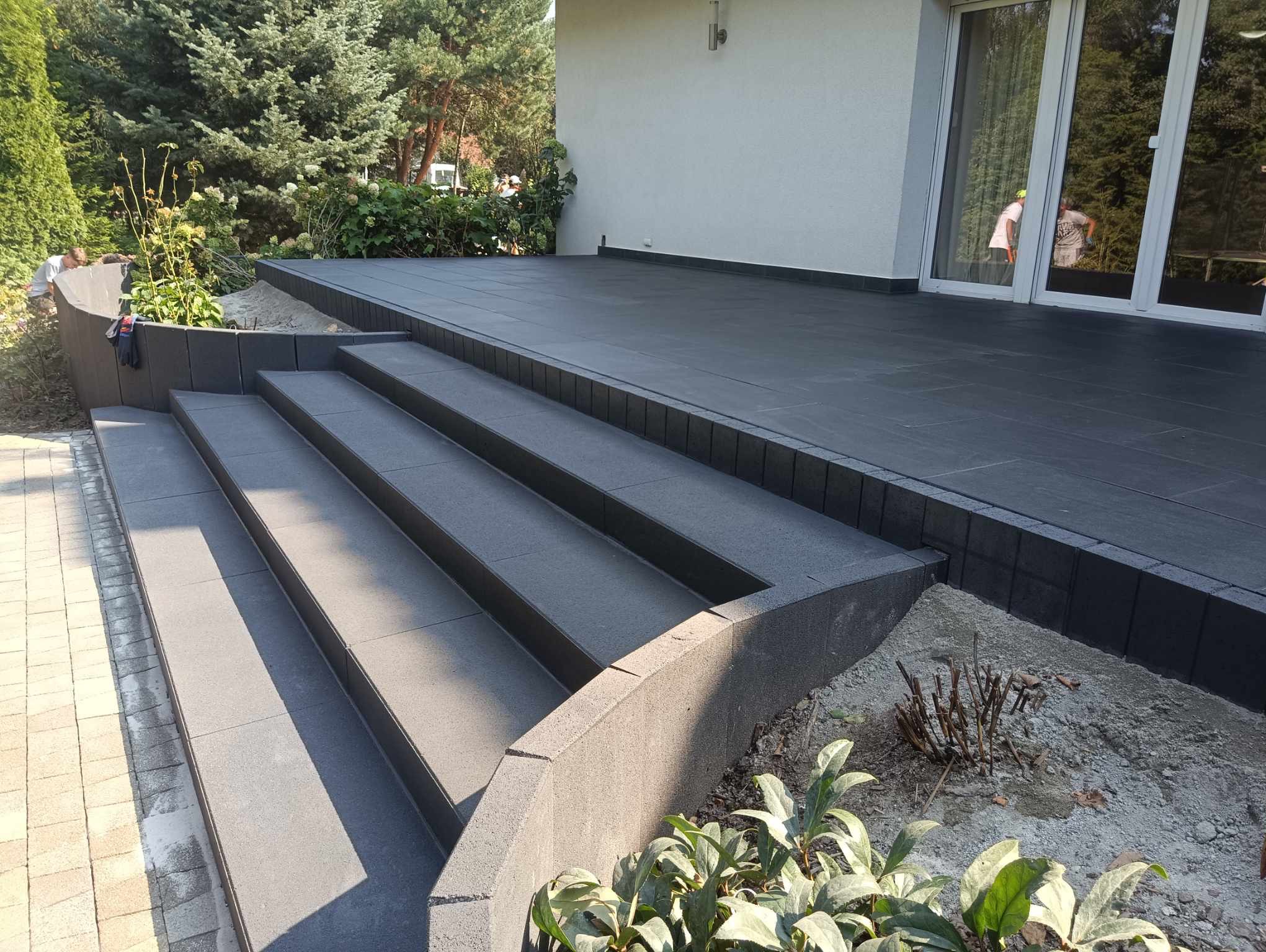 Nowoczesny taras z ciemnoszarej kostki, schody z tego samego materiału. Widoczne detale wykończenia i zieleń w tle. Ujęcie z niskiej perspektywy.