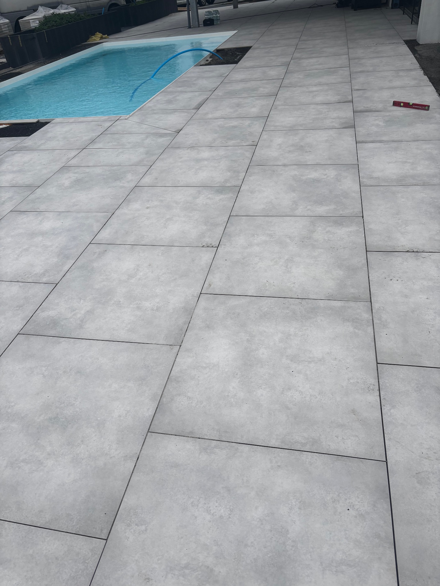 Jasnoszare płyty tarasowe ułożone wokół basenu, z widoczną poziomicą. Część płyt jeszcze nieułożona. Minimalistyczny design.