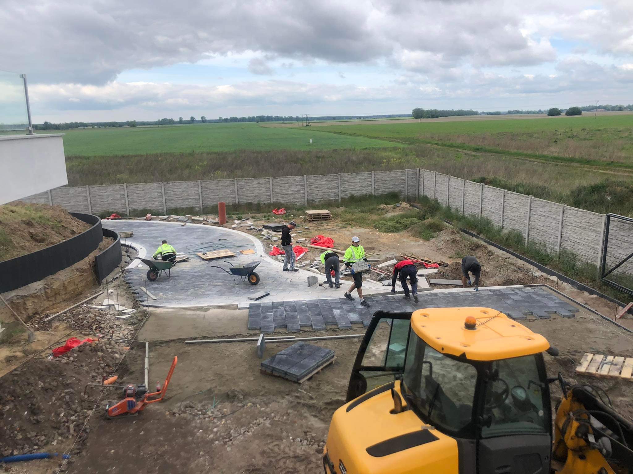 Ekipa brukarzy układa szarą kostkę na podjeździe. W tle widoczny krajobraz polny i maszyny budowlane, w tym żółty traktor. Praca w toku, dzień pochmurny.