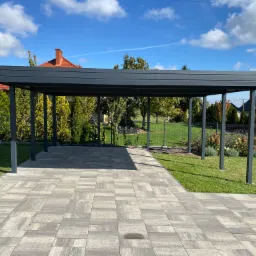 Nowoczesna, szara konstrukcja zadaszenia nad tarasem w Stobiernej, widok z przodu na kostkę brukową, w tle trawnik, ogrodzenie i drzewa pod błękitnym niebem.