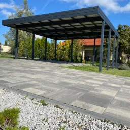 Nowoczesna, grafitowa konstrukcja zadaszenia na podjeździe z szarej kostki brukowej. W tle widoczny dom z czerwoną dachówką i zadbany ogród pod błękitnym niebem.