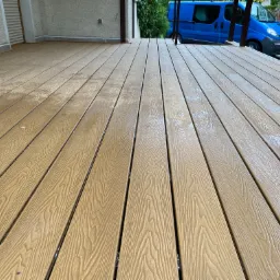 Nowy taras z desek kompozytowych w kolorze jasnego drewna, widok z niskiej perspektywy, w tle zaparkowany niebieski van.