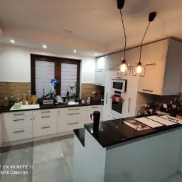 Nowoczesna kuchnia z białymi szafkami, czarnym blatem i wyspą, oświetlona wiszącymi lampami w stylu industrialnym, z elementami dekoracyjnymi i oknem z żaluzjami.