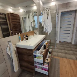 Nowoczesna łazienka z drewnianą szafką pod umywalką z otwartymi szufladami, widoczne kosmetyki i akcesoria, na ścianach beżowe płytki, w tle kabina prysznicowa i białe ręczniki.