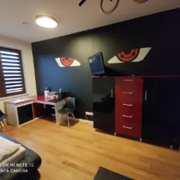 Pokój młodzieżowy z czarną ścianą ozdobioną malunkami oczu, biurkiem z czerwonym blatem, szafką RTV w kolorach czerni i czerwieni oraz drewnianą podłogą.