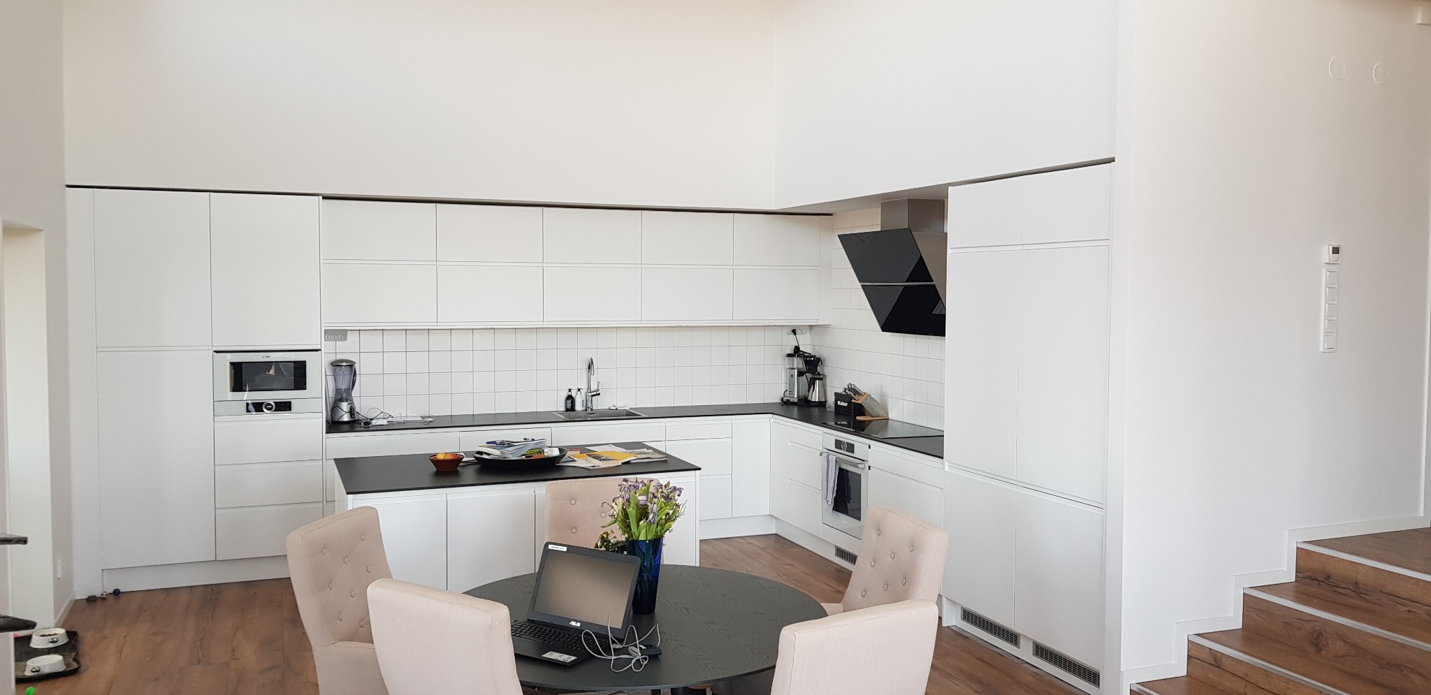 Jasna kuchnia z białymi, matowymi szafkami, czarnym blatem i wyspą, okrągłym stołem z laptopem i kwiatami, oraz schodami z drewnianymi stopniami w tle.