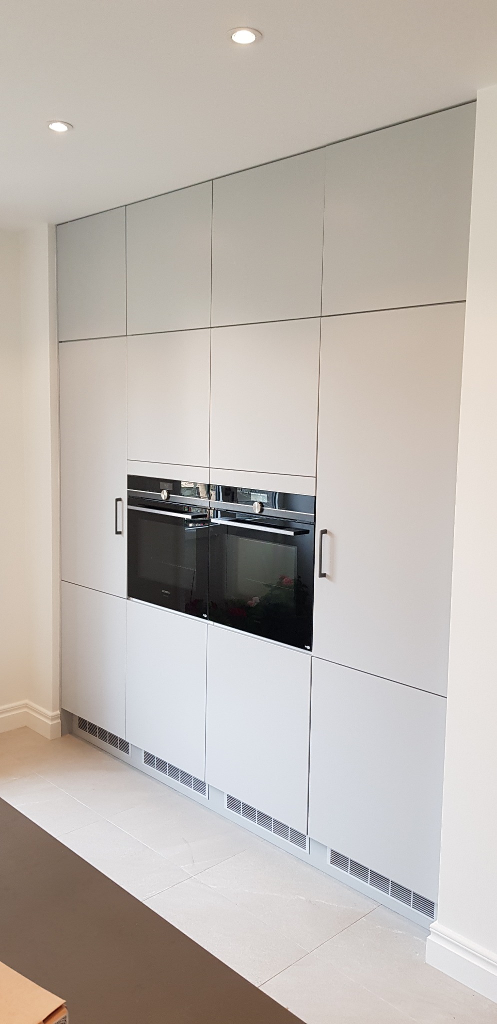 Nowoczesna zabudowa kuchenna w odcieniach szarości z dwoma piekarnikami Siemens, minimalistyczne uchwyty, oświetlenie punktowe w suficie i kratki wentylacyjne u dołu szafek.