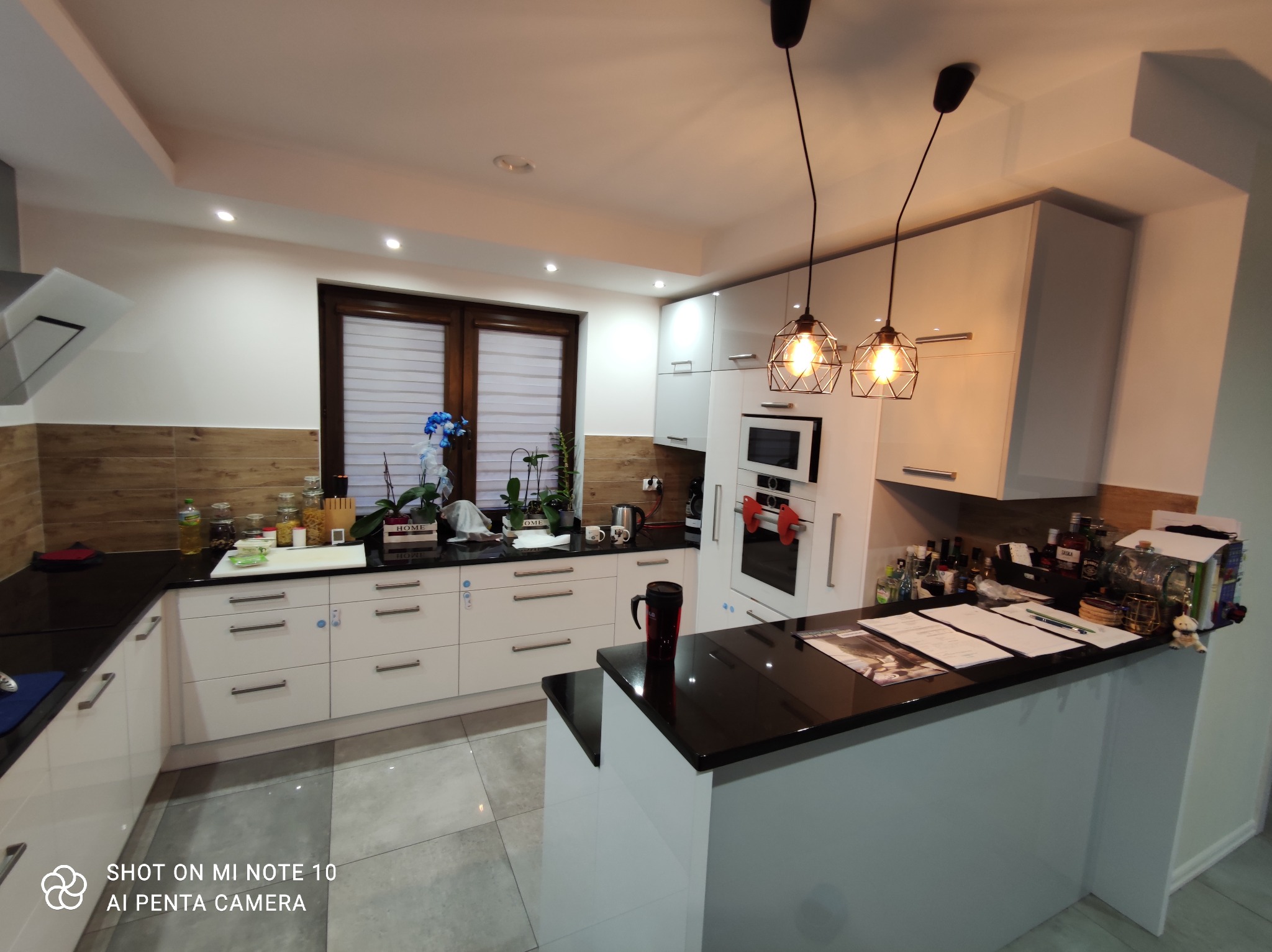 Nowoczesna kuchnia z białymi szafkami, czarnym blatem i wyspą, oświetlona wiszącymi lampami w stylu industrialnym, z elementami dekoracyjnymi i oknem z żaluzjami.