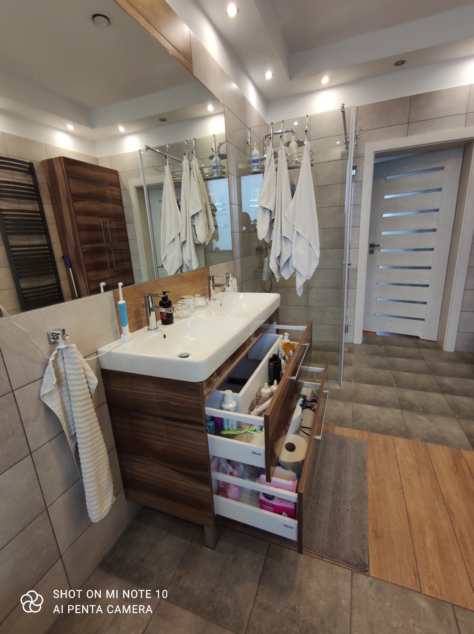 Nowoczesna łazienka z drewnianą szafką pod umywalką z otwartymi szufladami, widoczne kosmetyki i akcesoria, na ścianach beżowe płytki, w tle kabina prysznicowa i białe ręczniki.