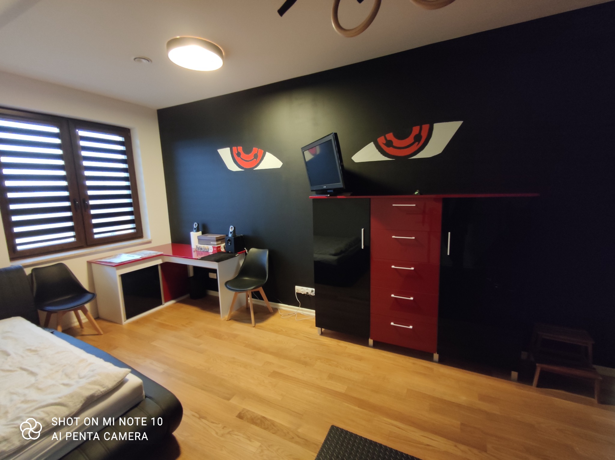 Pokój młodzieżowy z czarną ścianą ozdobioną malunkami oczu, biurkiem z czerwonym blatem, szafką RTV w kolorach czerni i czerwieni oraz drewnianą podłogą.