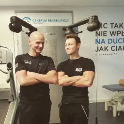 Dwaj mężczyźni w czarnych koszulkach z logo, stojący na tle sali rehabilitacyjnej z bieżnią i urządzeniem do rehabilitacji Technogym, z uśmiechami na twarzach i skrzyżowanymi ramionami.