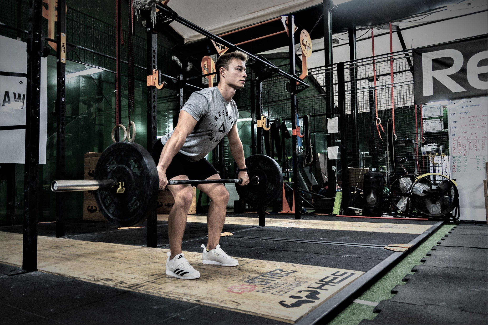 Młody mężczyzna w szarej koszulce z krótkim rękawem i czarnych spodenkach podnosi sztangę w siłowni; widoczne wyposażenie siłowni, drabinki, podłoga z maty gumowej i drewniany podest pod sztangą.
