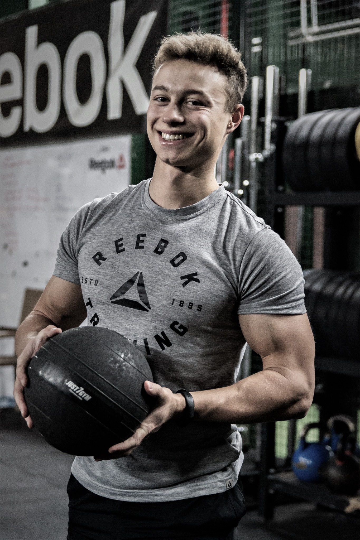 Uśmiechnięty młody mężczyzna w szarej koszulce Reebok trzyma czarną piłkę lekarską Just7Gym na tle siłowni z obciążeniami i logo Reebok.