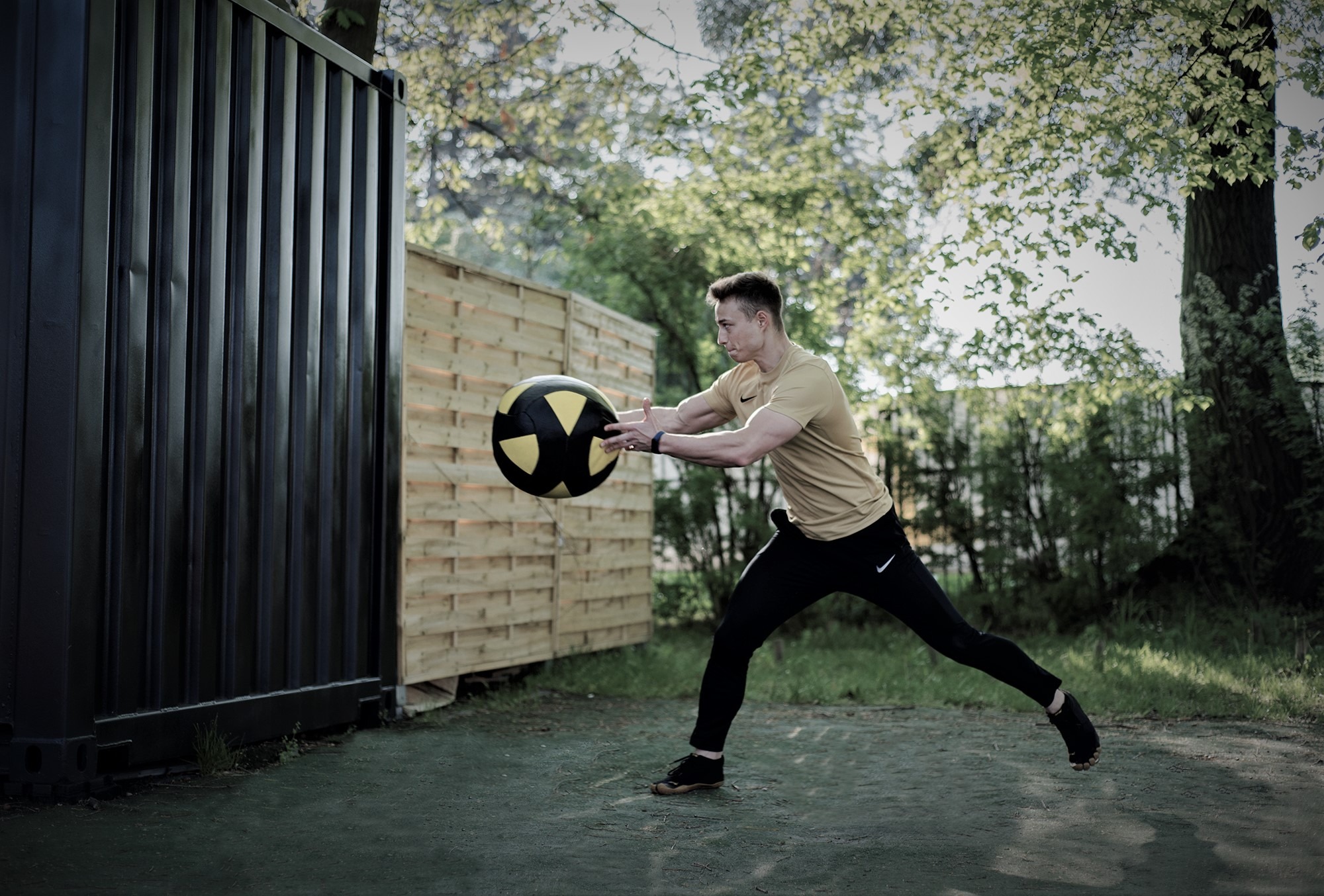 Młody mężczyzna w sportowym stroju ćwiczy z czarno-żółtą piłką lekarską na tle kontenera i drewnianego płotu, w otoczeniu zieleni.