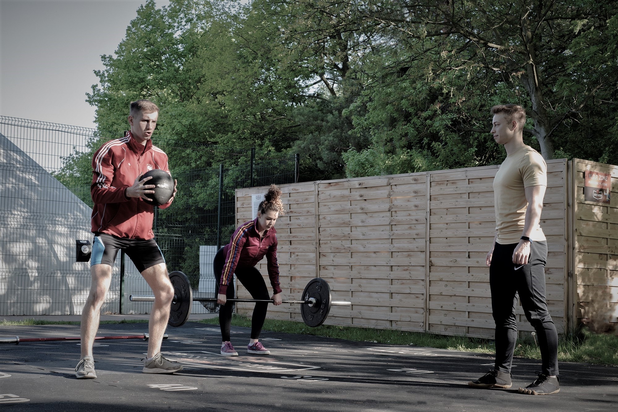 Troje osób ćwiczy na zewnątrz: mężczyzna trzyma piłkę lekarską, kobieta podnosi sztangę, a mężczyzna obserwuje z boku, na tle drewnianego ogrodzenia i zieleni.