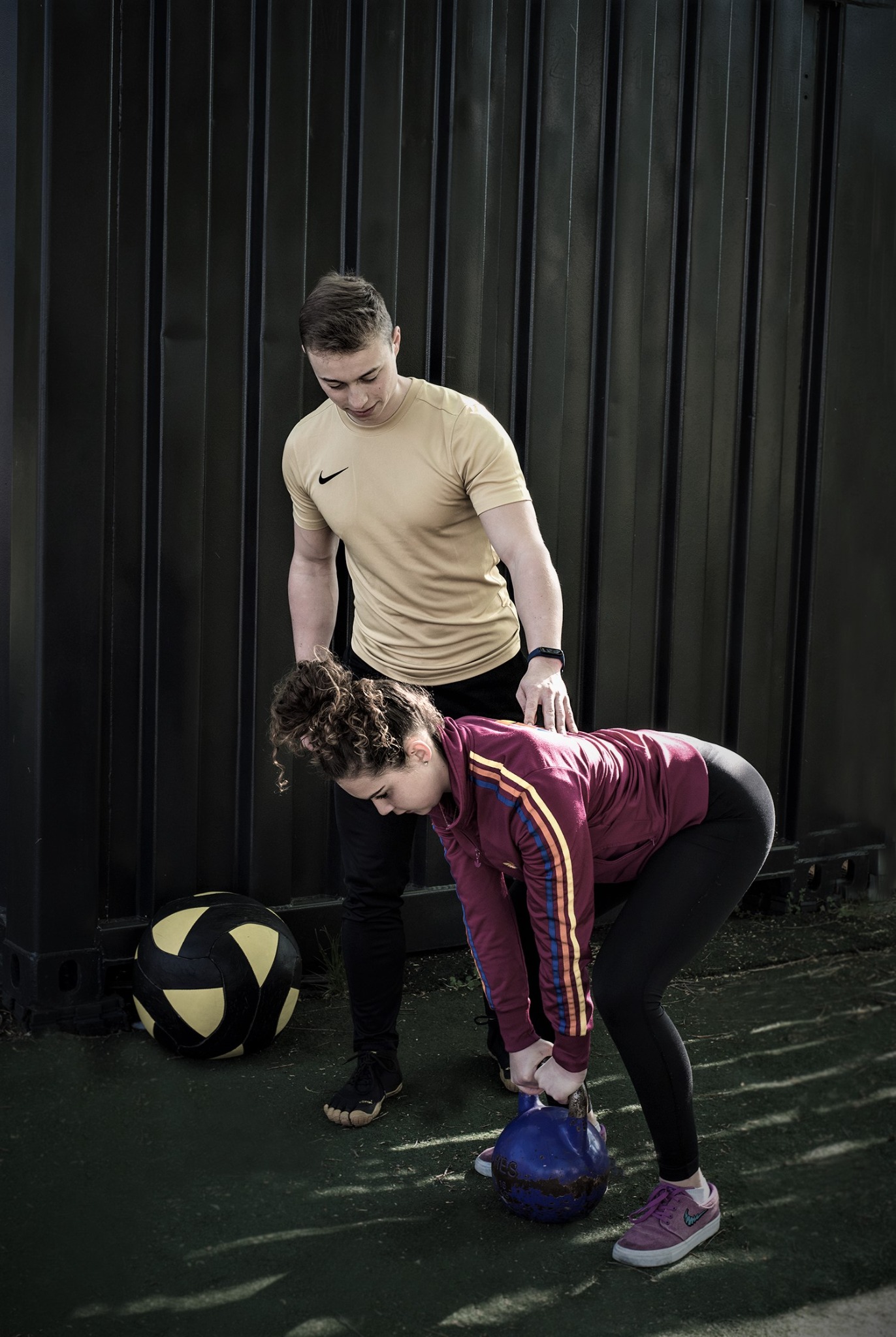 Trener personalny asekuruje kobietę podczas podnoszenia niebieskiego kettlebella w plenerowej siłowni; w tle czarny, metalowy kontener i piłka do ćwiczeń.