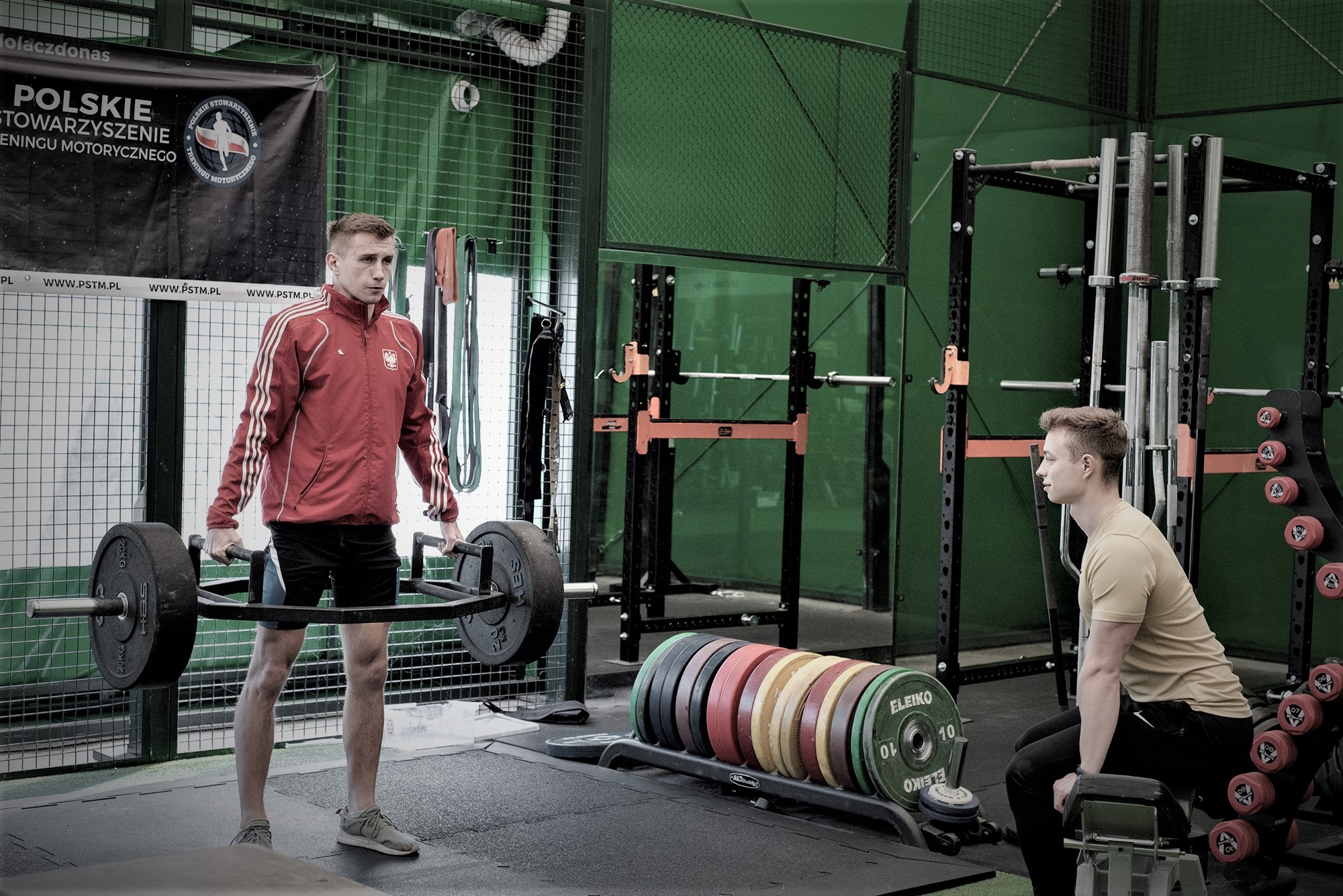 Młody mężczyzna w czerwonej bluzie z godłem Polski podnosi sztangę typu trap bar w siłowni; w tle widoczny drugi mężczyzna siedzący na ławce i sprzęt do ćwiczeń.