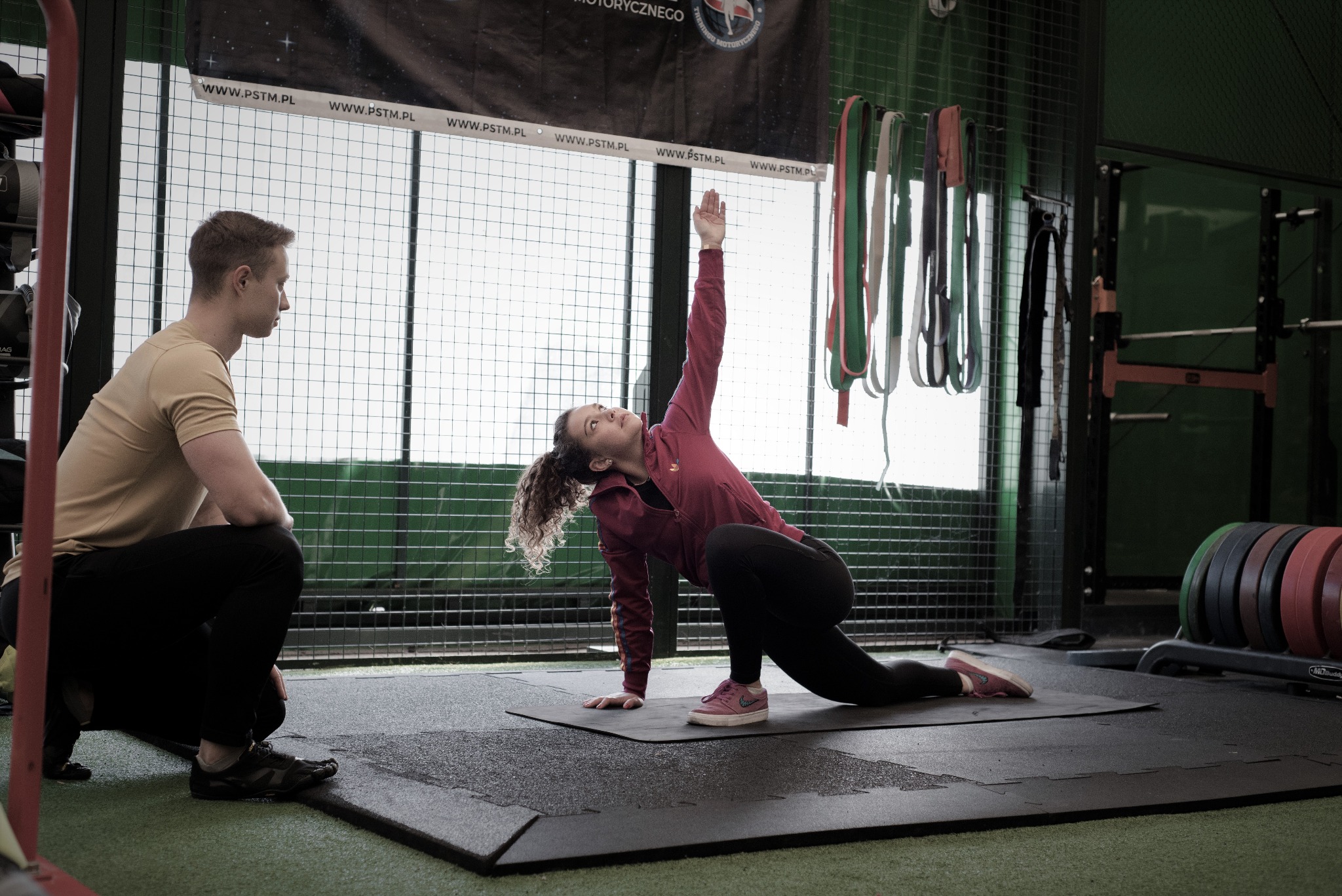 Młoda kobieta w różowej bluzie wykonuje ćwiczenie rozciągające na macie w siłowni, pod okiem trenera, który przykuca obok; w tle widoczne obciążenia i drabinki.