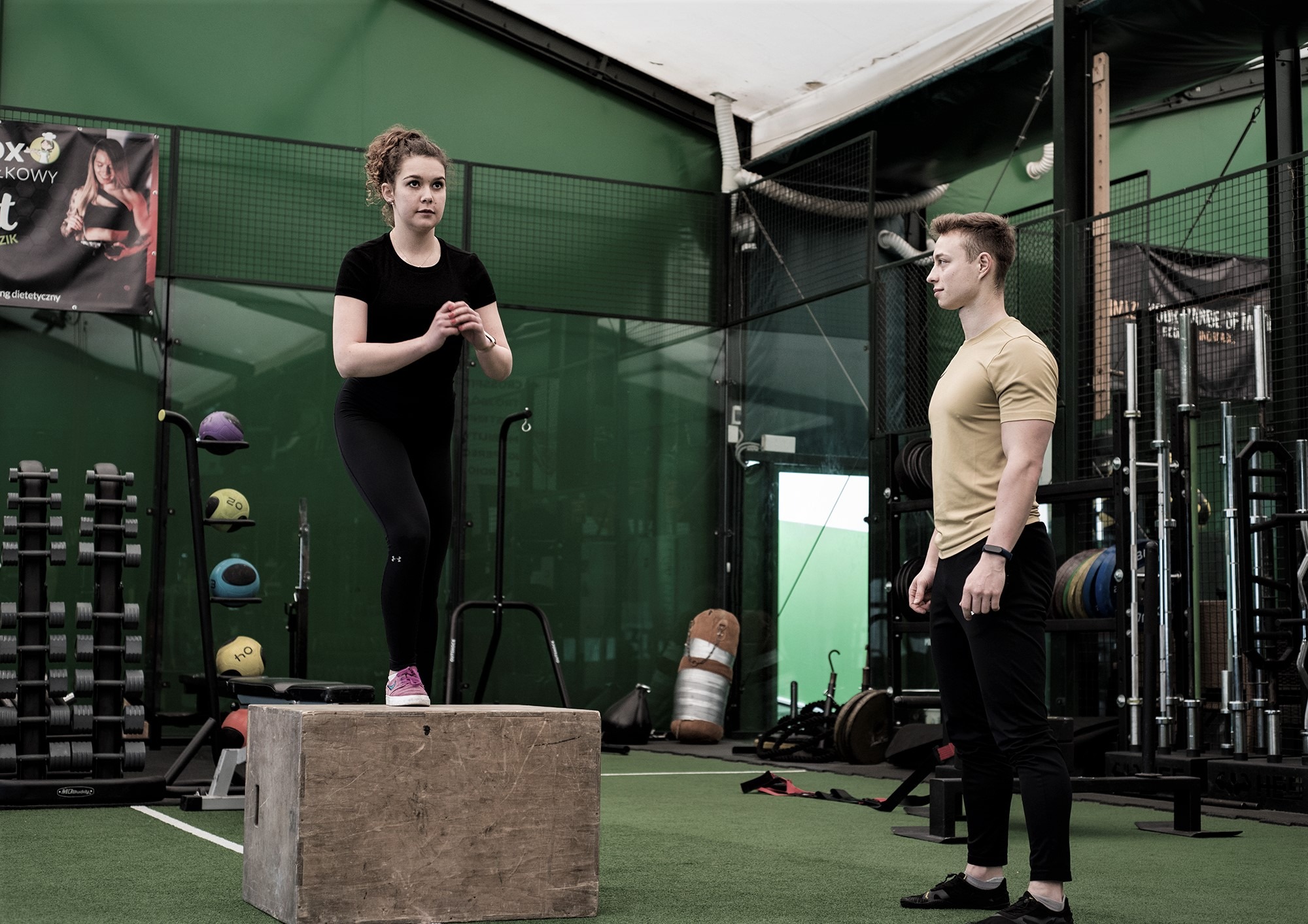 Kobieta w czarnym stroju sportowym wykonuje ćwiczenie wskakiwania na drewnianą skrzynię pod nadzorem mężczyzny w żółtej koszulce, w tle sprzęt do ćwiczeń i reklama siłowni.