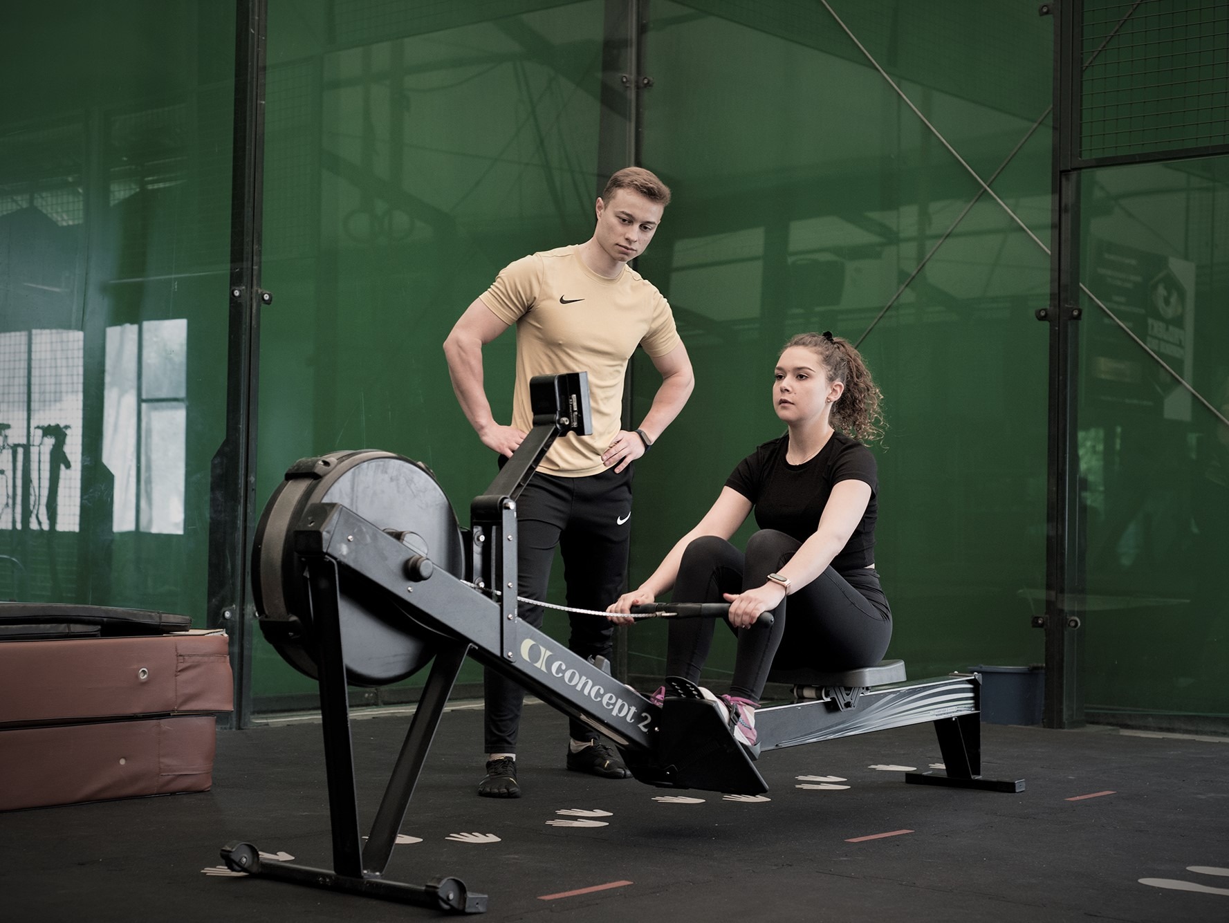 Kobieta ćwiczy na ergometrze wioślarskim Concept2, nadzorowana przez mężczyznę w sportowym stroju, na tle zielonej ściany w siłowni.