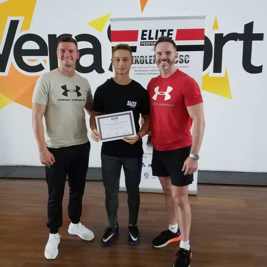 Trzech mężczyzn pozuje do zdjęcia: dwóch w sportowych koszulkach Under Armour, jeden trzyma certyfikat Elite Performance.