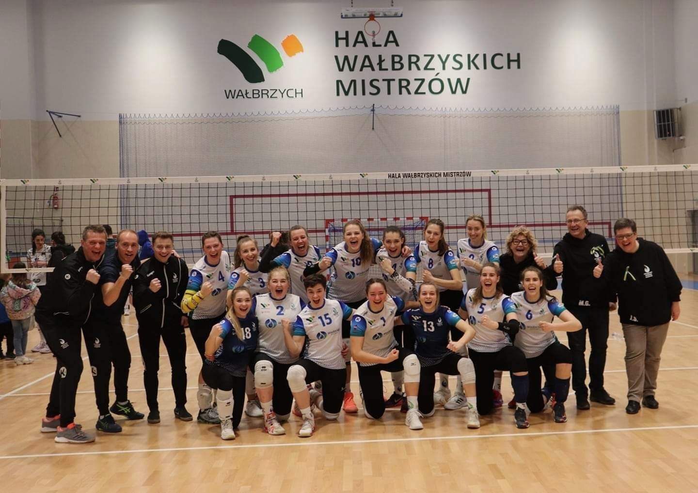 Drużyna siatkarska świętuje zwycięstwo na tle siatki i napisu Hala Wałbrzyskich Mistrzów, zawodniczki w sportowych strojach z numerami, trenerzy i kibice z podniesionymi pięściami w geście triumfu.