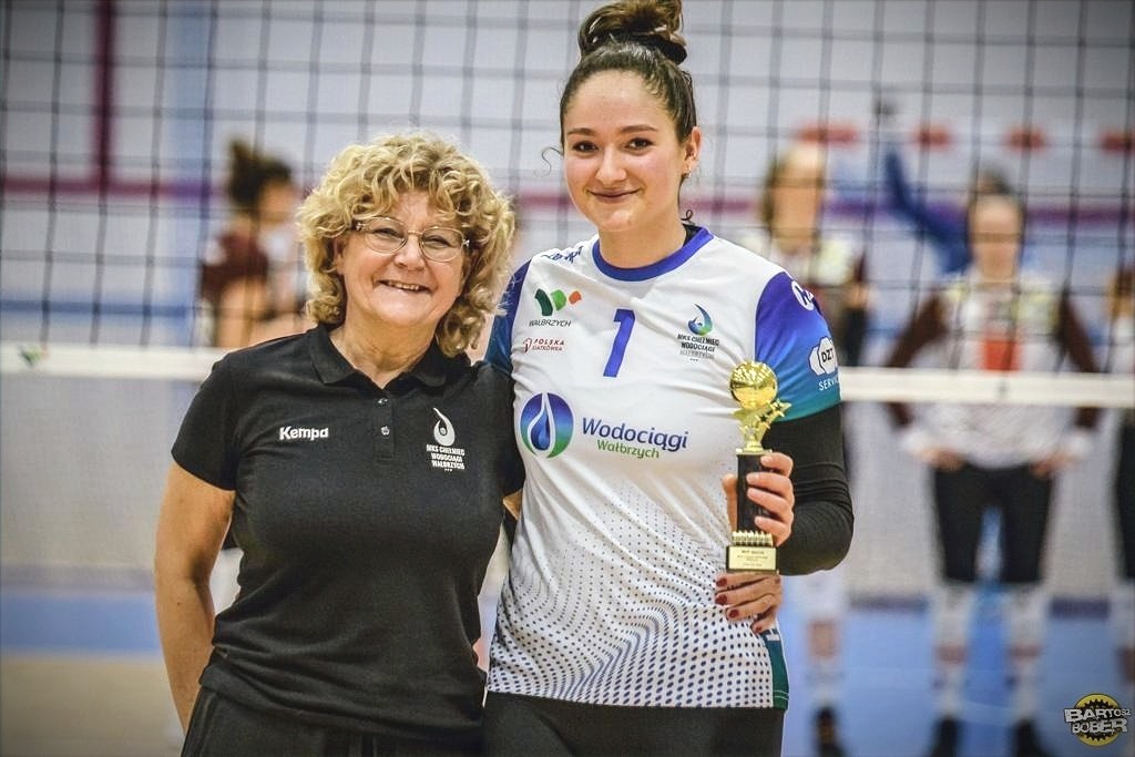 Uśmiechnięta siatkarka w koszulce z numerem 1 i logo Wodociągów Wałbrzych trzyma złote trofeum, obok niej stoi trenerka w czarnej koszulce z logo klubu, w tle siatka i inni zawodnicy.