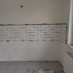 Ściana z białymi, prostokątnymi płytkami ułożonymi w cegiełkę, częściowo wykończona, widoczne rury instalacji wodnej i gniazdko elektryczne, surowy stan wykończenia.