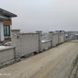 Nowoczesny, modułowy płot z szarej cegły klinkierowej z czarnymi metalowymi uchwytami, oddzielający posesję od drogi, w tle dom z szarą dachówką i zaśnieżone pole w pochmurny dzień.