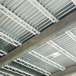 CONSTATICA Pracownia Projektowa - Metalowy strop z białymi, kratownicowymi wzmocnieniami i betonowymi belkami, widok z dołu.