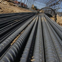 CONSTATICA Pracownia Projektowa - Stalowe pręty zbrojeniowe ułożone równolegle na placu budowy, w tle widoczna konstrukcja stalowa i maszyna budowlana.