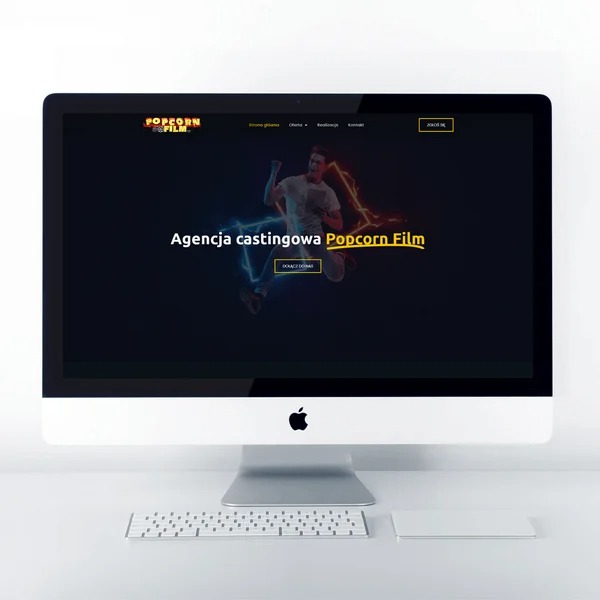 Ekran komputera iMac wyświetlający stronę internetową agencji castingowej Popcorn Film z dynamiczną grafiką postaci w locie otoczonej efektami świetlnymi, na białym biurku z klawiaturą i Magic...