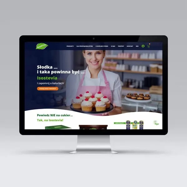 Monitor wyświetlający stronę internetową Isostevia z uśmiechniętą kobietą w różowym fartuchu trzymającą talerz z babeczkami z kremem i wiśniami, w tle regały z produktami.