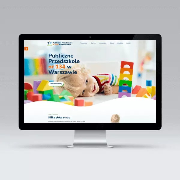 Monitor wyświetlający stronę internetową przedszkola 'Publiczne Przedszkole nr 134 w Warszawie' z uśmiechniętym dzieckiem i kolorowymi klockami na banerze.