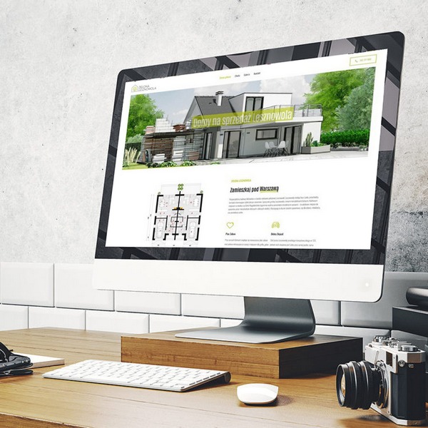 Ekran monitora wyświetlający stronę internetową z ofertą sprzedaży domu w Lesznowoli, widoczny plan domu, klawiatura, mysz i aparat fotograficzny na drewnianym biurku, tło z białych płytek.