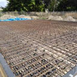 Zbrojenie płyty fundamentowej z dystansami, przygotowane do zalania betonem, widok z góry na plac budowy z ogrodzeniem z siatki.