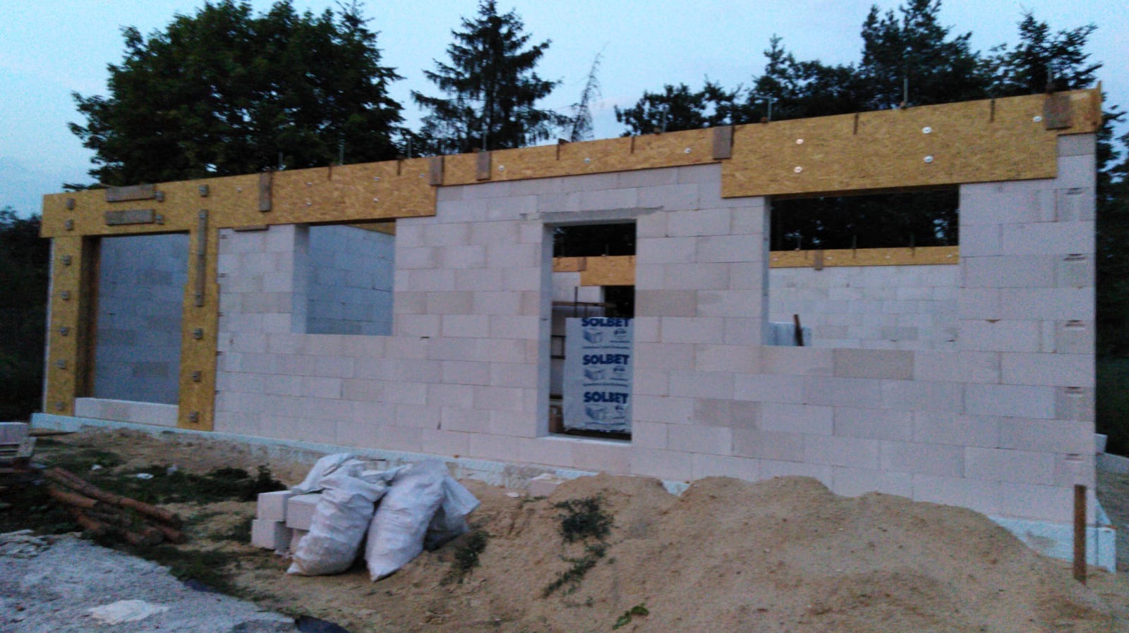 Wznoszenie ścian z betonu komórkowego, widoczne otwory okienne i drzwiowe z drewnianymi nadprożami i elementami szalunkowymi, worki z zaprawą murarską u podstawy ściany, w tle drzewa.