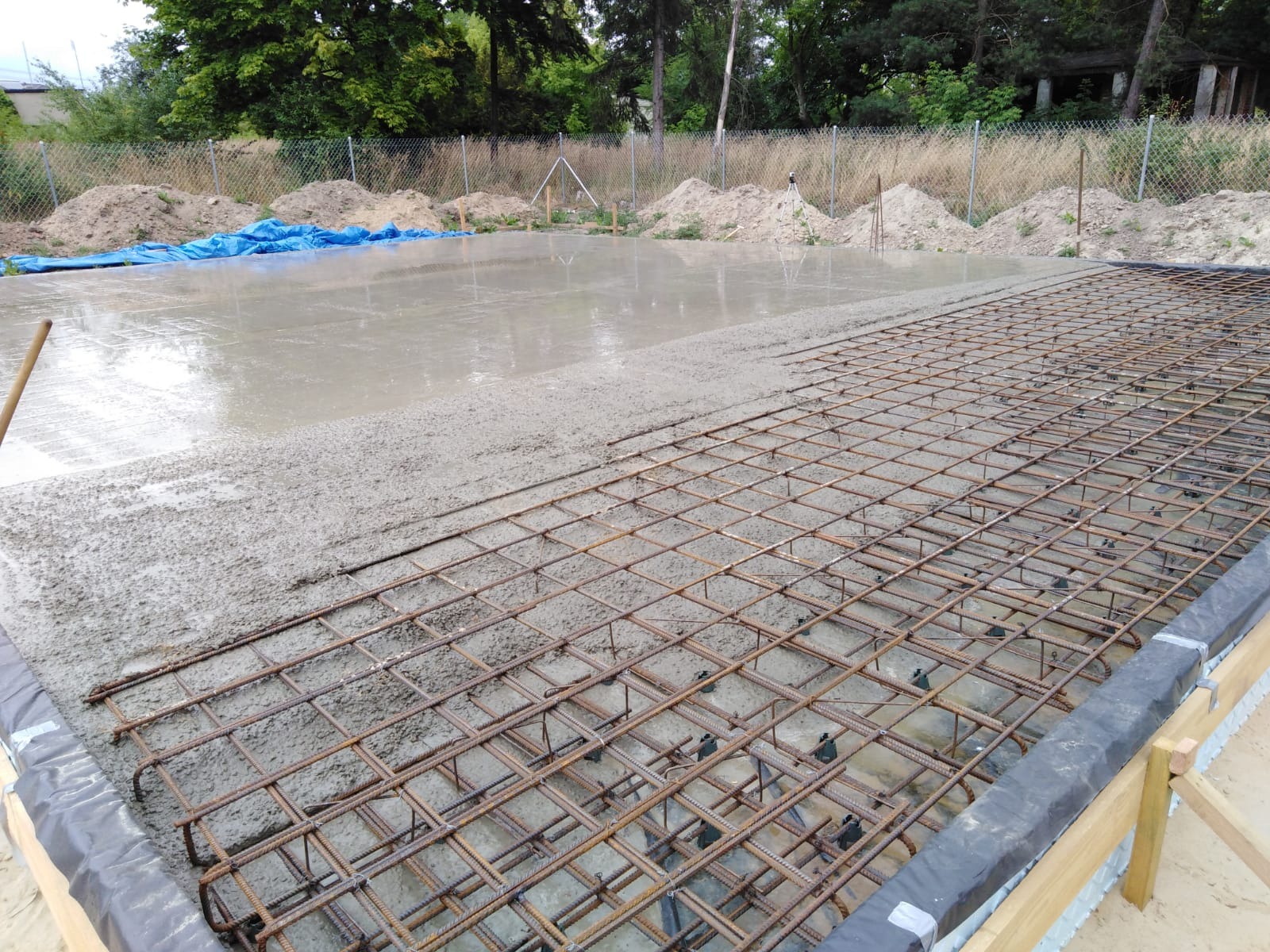 Wylewanie betonu na zbrojenie fundamentu, widoczne szalunki i wystające pręty zbrojeniowe, w tle pryzmy ziemi i ogrodzenie z siatki.