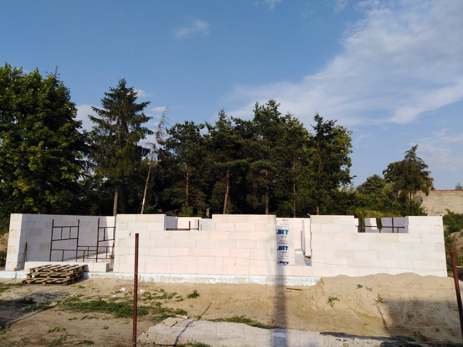 Wznoszone ściany parteru z betonu komórkowego na fundamencie, widoczne zbrojenia i izolacja, w tle drzewa i błękitne niebo.