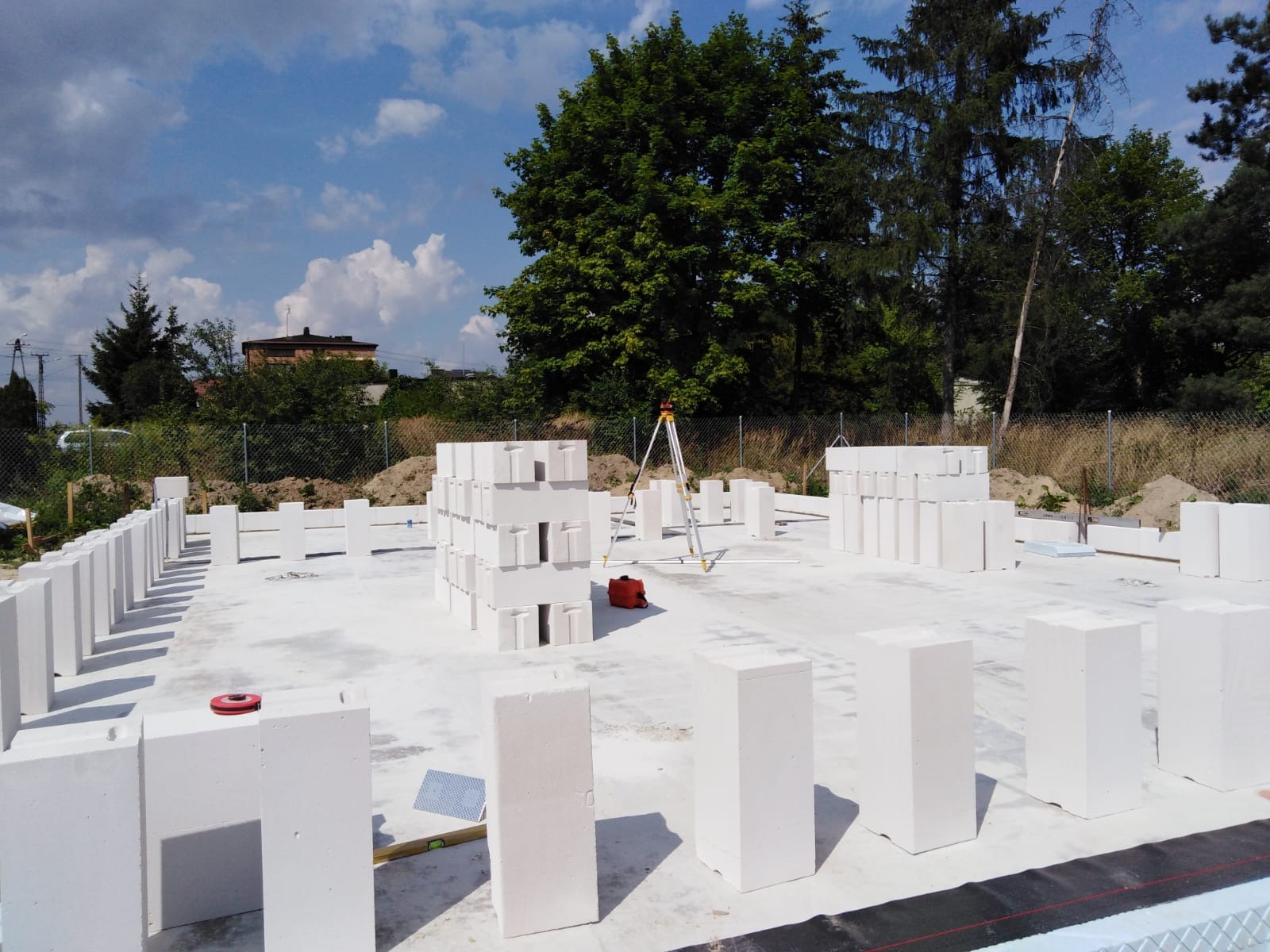 Wznoszenie ścian domu z bloczków betonu komórkowego na przygotowanej płycie fundamentowej, widoczny niwelator laserowy i poziomica.