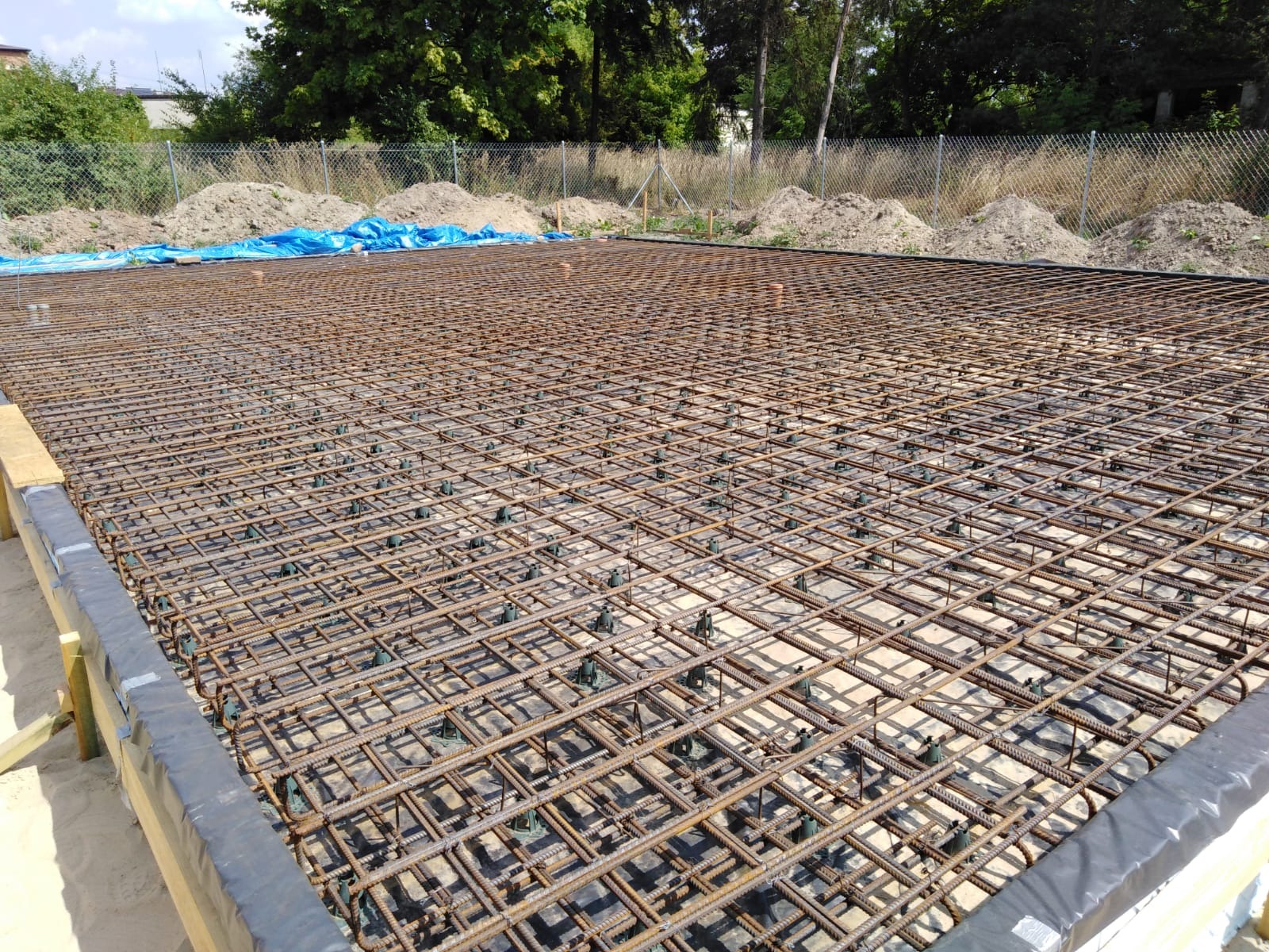 Zbrojenie płyty fundamentowej z dystansami, przygotowane do zalania betonem, widok z góry na plac budowy z ogrodzeniem z siatki.