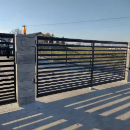 Nowoczesna, pozioma brama wjazdowa z czarnymi, płaskimi profilami i słupkami z szarej kostki, wyposażona w domofon i lampę ostrzegawczą, na tle brukowanej nawierzchni i pola.